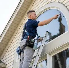 Professional window cleaner on a ladder using a squeegee to clean an exterior arched window, Henderson, NV.
